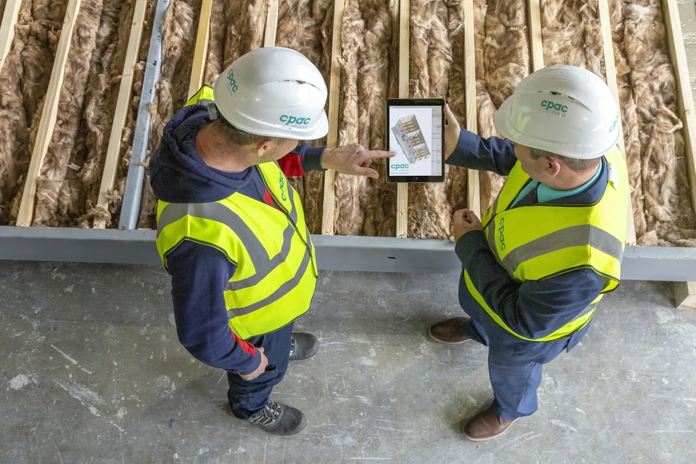 Two construction workers in hard hats and safety vests reviewing plans on a tablet